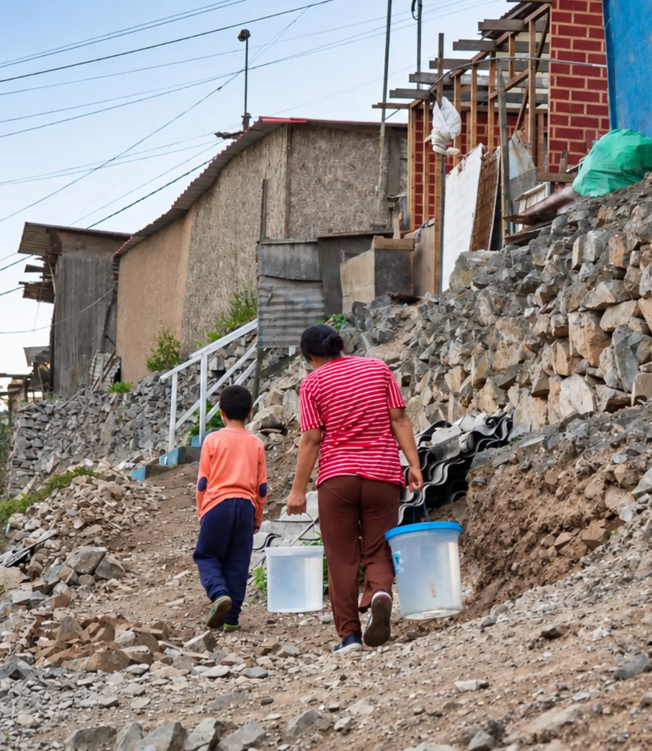 A mother and her son carrying buckets of water up a rocky hillside path in a Lima community